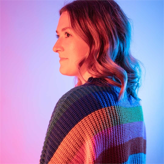 A student smiling off to the side of the camera, their back is turned and they're wearing a colourful knitted jumper