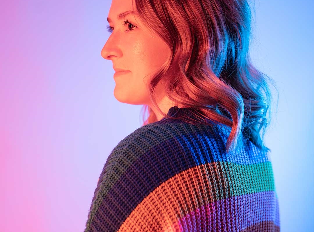 A student smiling off to the side of the camera, their back is turned and they're wearing a colourful knitted jumper