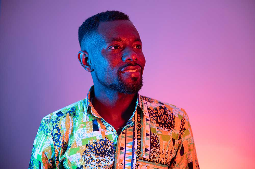 A photograph of Emmanuel Owusu Takyi wearing a colourful patterned shirt, lit up by red and blue lights