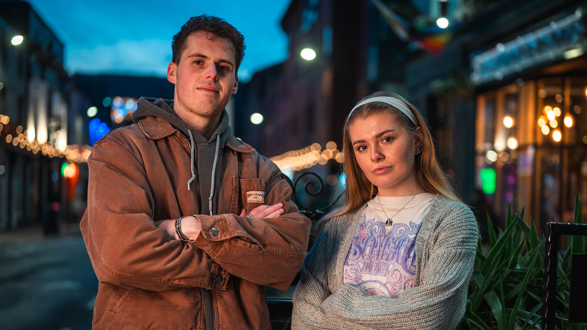 Two students standing outdoors at dusk, looking at the camera with arms crossed