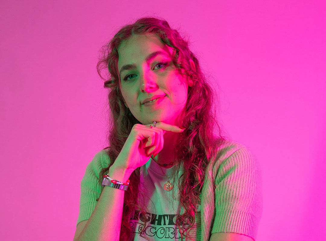 Student with their chin resting on their hand, smiling gently while stood in front of a pink backdrop.