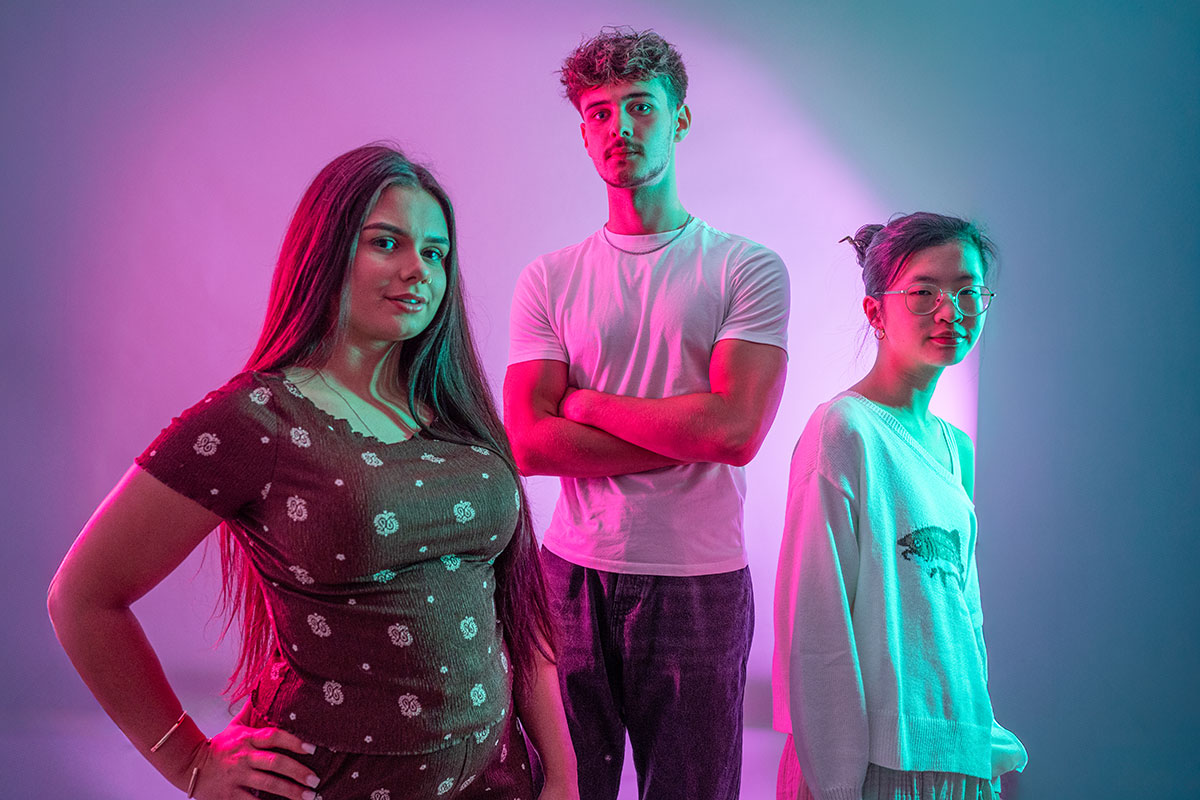 Three students standing looking towards the camera, lit with purple lights.