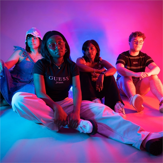 Four students sat on the floor looking at the camera, lit up by neon pink lights