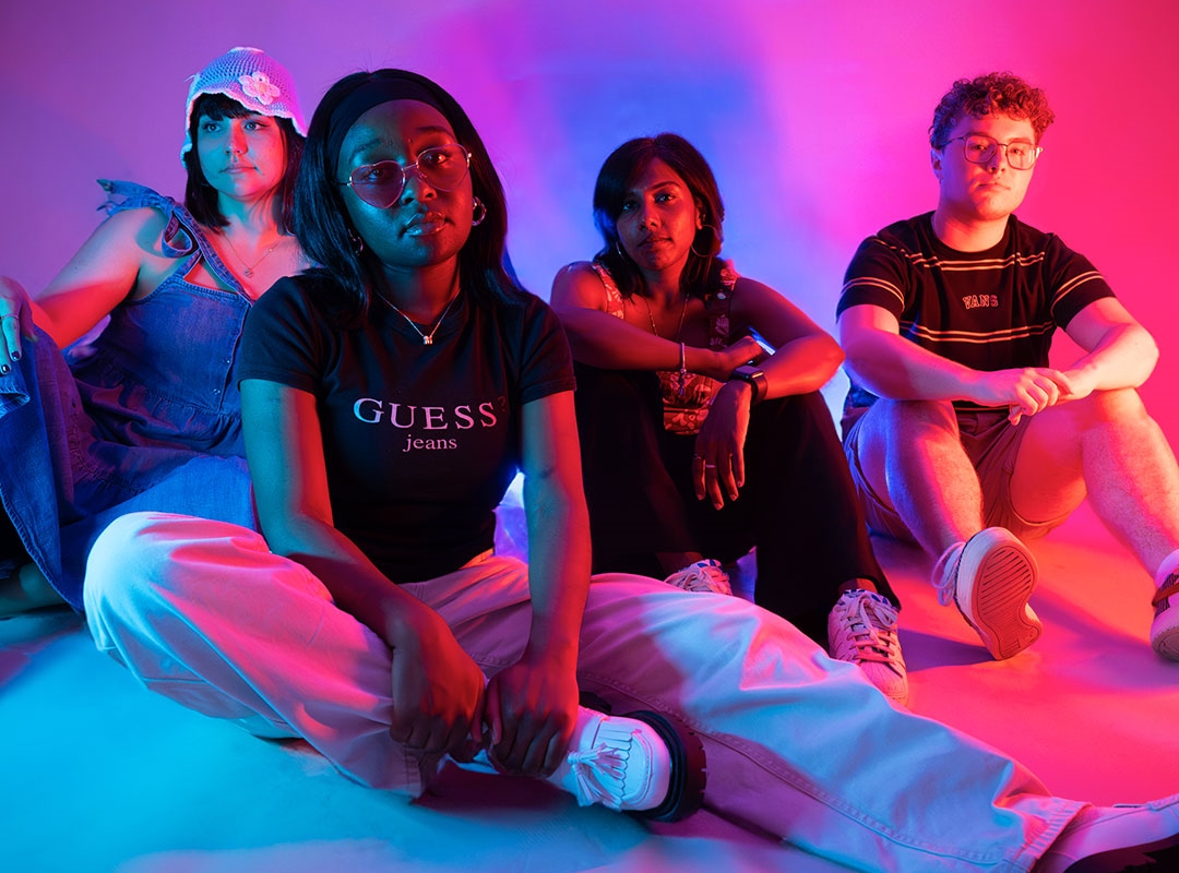 Four students sat on the floor looking at the camera, lit up by neon pink lights