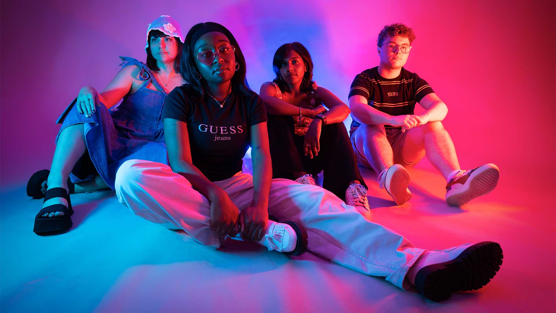 Four students sat on the floor in a photography studio, light up by neon pink and purple lights