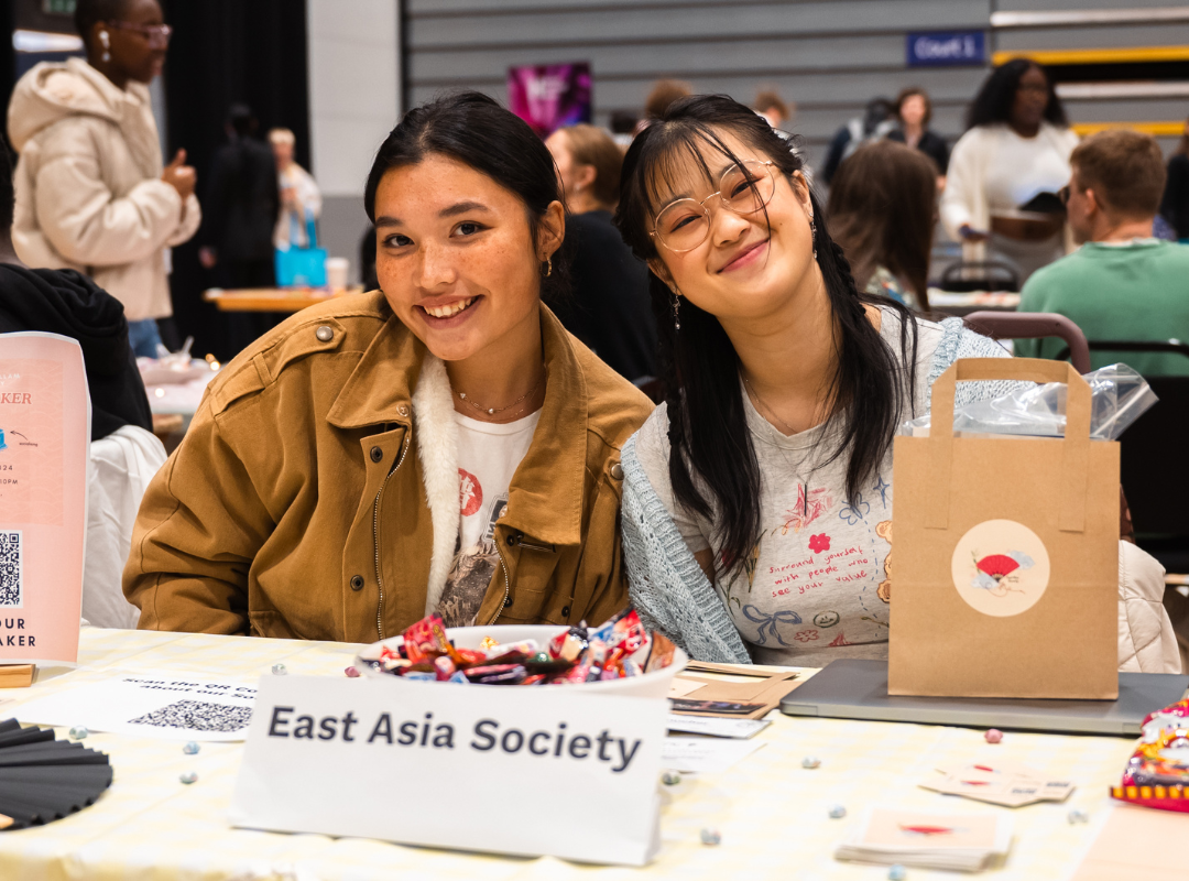 Two smiling students at the 'East Asia Society' table during a university event, with sweets, flyers, and a gift bag on display.