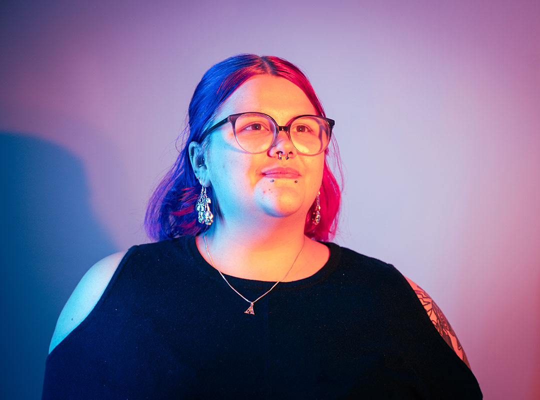 Student with glasses and piercings looking into the distance, lit with a glow of blue and pink lights