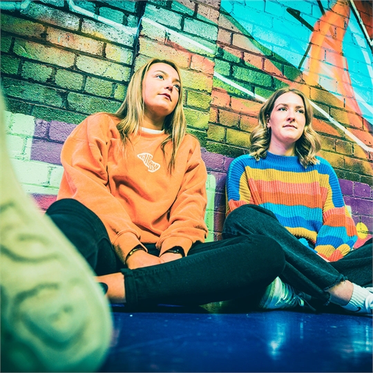 Two students leaning against a colourful graffiti wall