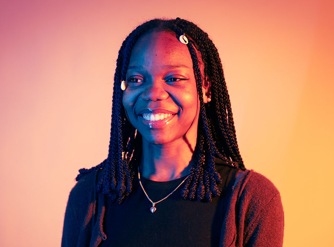 Student looking into distance and smiling. They're lit behind with soft orange and pink lighting.