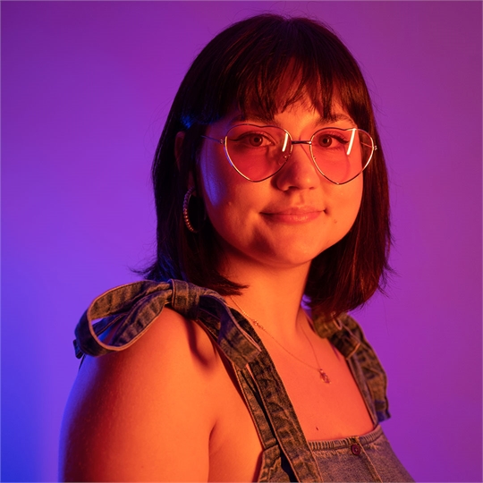 A student wearing dungarees and love heart shaped glasses, lit up by neon lights