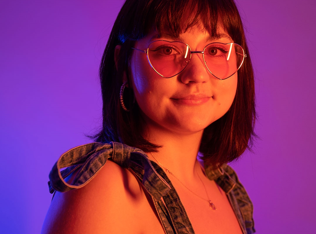 A student wearing dungarees and love heart shaped glasses, lit up by neon lights
