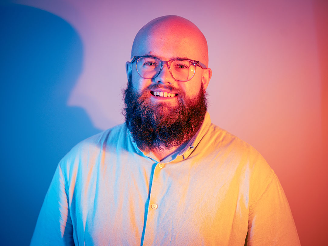 A student smiling at the camera, lit behind by orange and blue lights