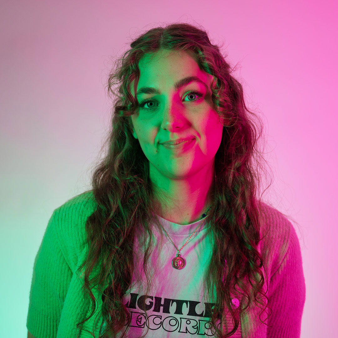 Molly Pemberton smiling at the camera and lit by bright green and pink lights.