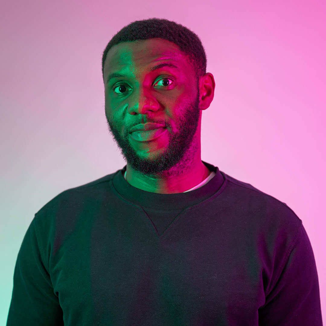 Ebuka H Obi looking towards the camera and lit by bright green and pink lights.