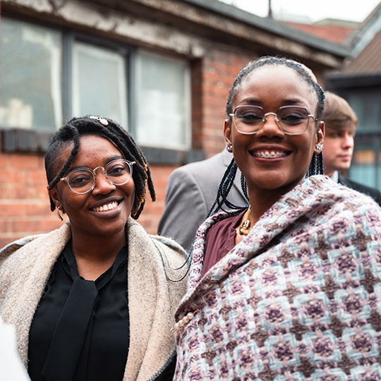 Two students, both smiling and wearing glasses, stand outdoors in front of a building.