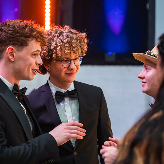 Three students wearing formal attire, suits and ties, engage in lively discussion.