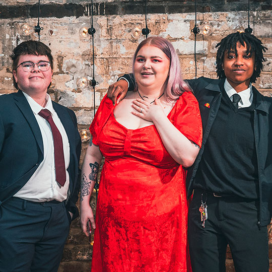 Three students wearing formal attire, one in a red dress and two in suits and ties, smile as they stand in front of a rustic brick wall.