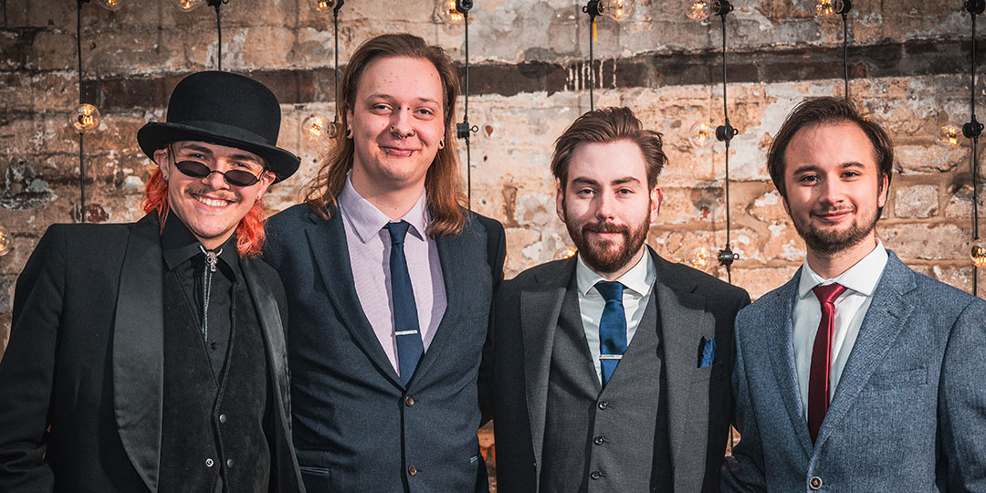 Four students wearing formal suits and ties smile as they pose for a group photo in front of a rustic brick wall.