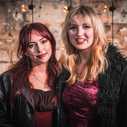 Two students wearing elegant formal dress smile as they stand side by side in front of a rustic brick wall.