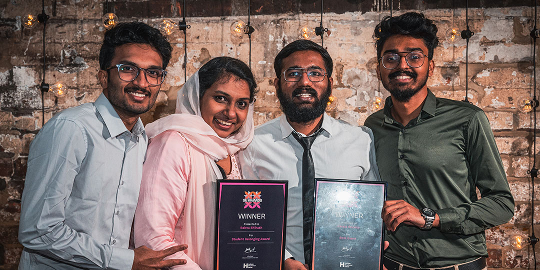 Four students proudly smile and display their awards as they stand in front of a rustic brick wall.