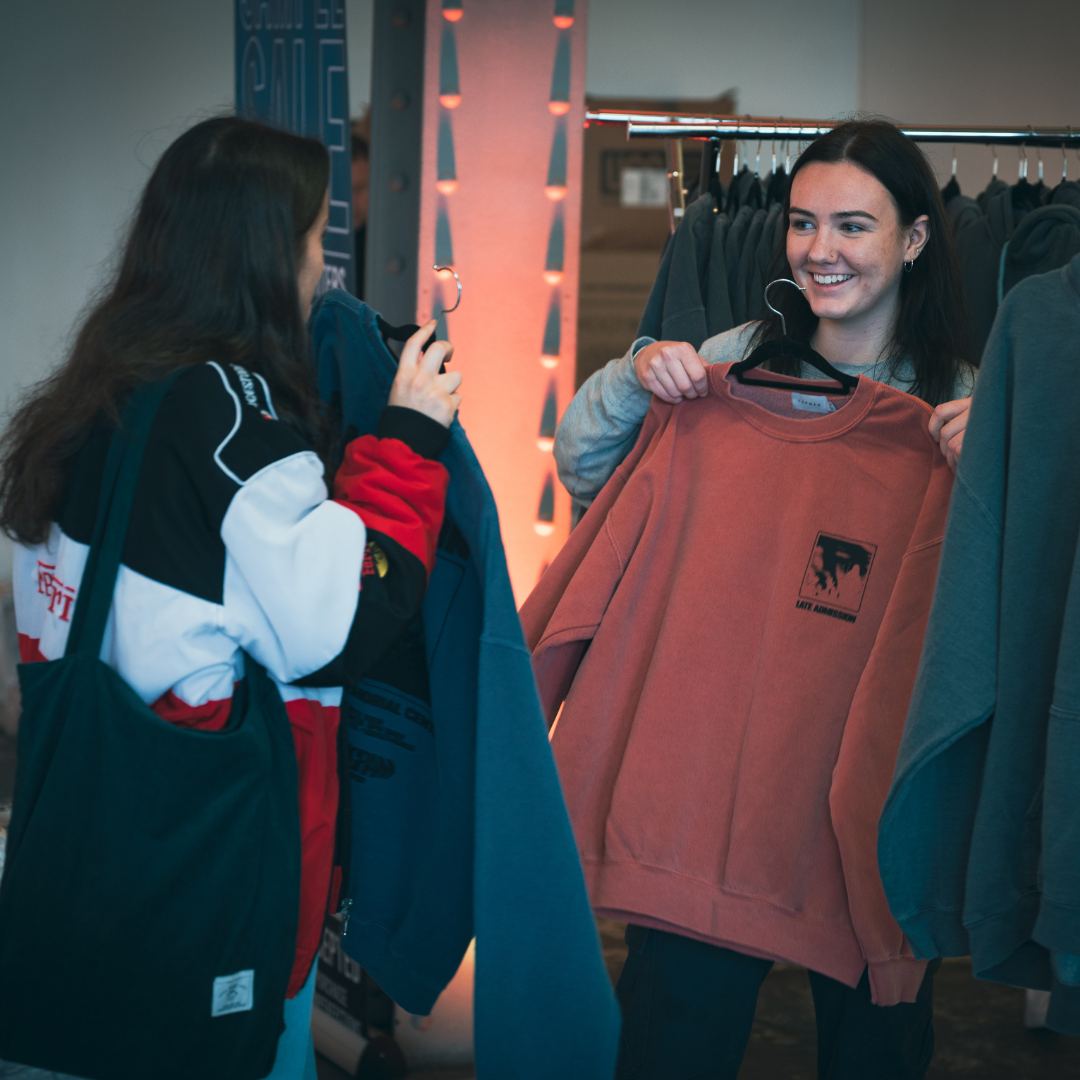 Two young female students smiling and holding up sweatshirts while organising clothing at a clothing rack.