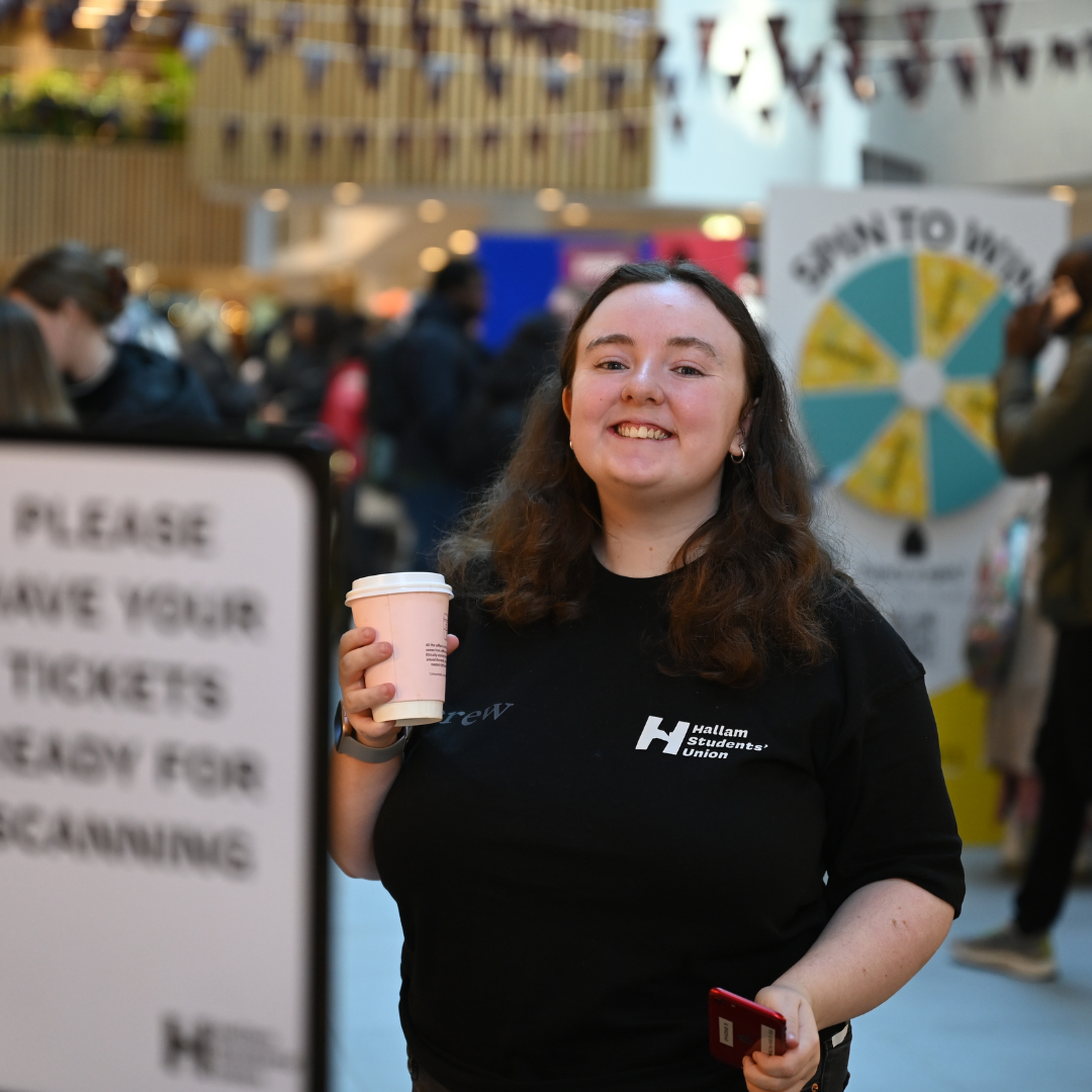 A Hallam Students' Union staff member smiles while holding a cup of coffee.