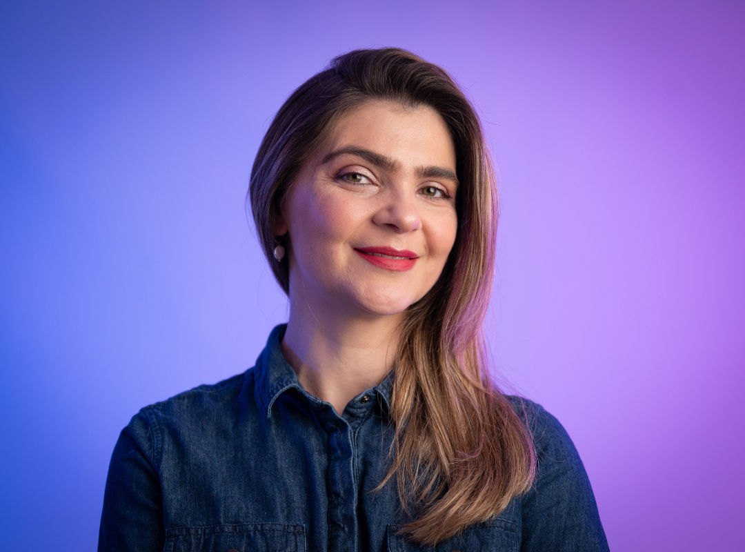A woman wearing a denim shirt and red lipstick, smiling confidently at the camera.