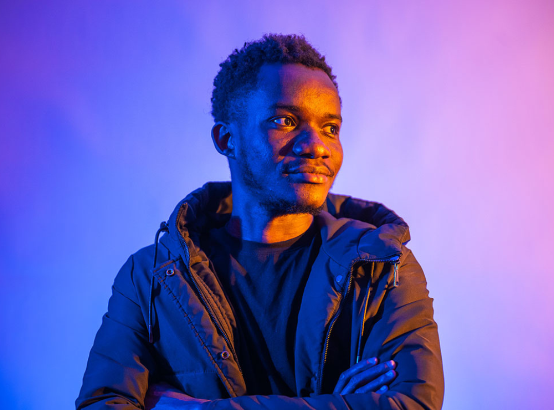Student lit with bright purple light looking towards the camera with a neutral expression. One arm is folded across their front and the other is bent with their hand resting on their ear.