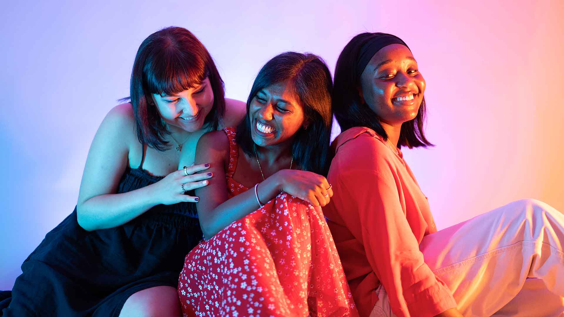 Three students sat on the floor laughing, lit up by neon orange and pink lighting