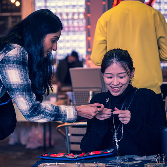 A student leaning over a table assisting a smiling student making jewellery at a Makers Market.