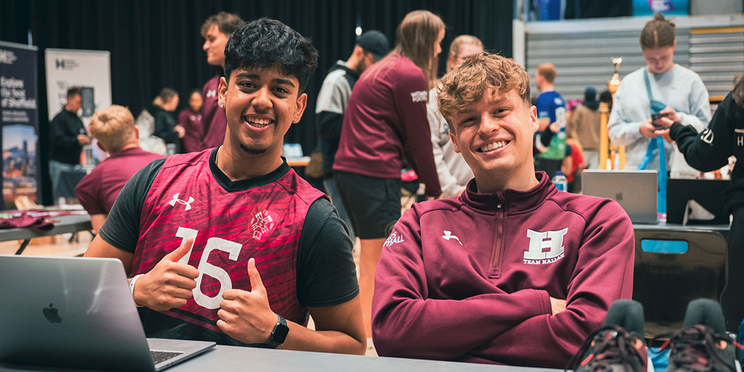 Two students in Hallam sports kit smiling as they sit behind a stall at the Societies and Sports Fair.