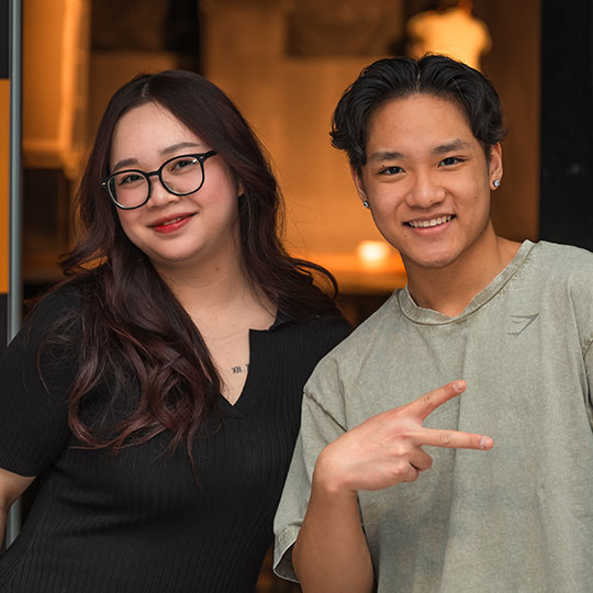 Two students smiling; one of them is making a sideways peace sign with their fingers.